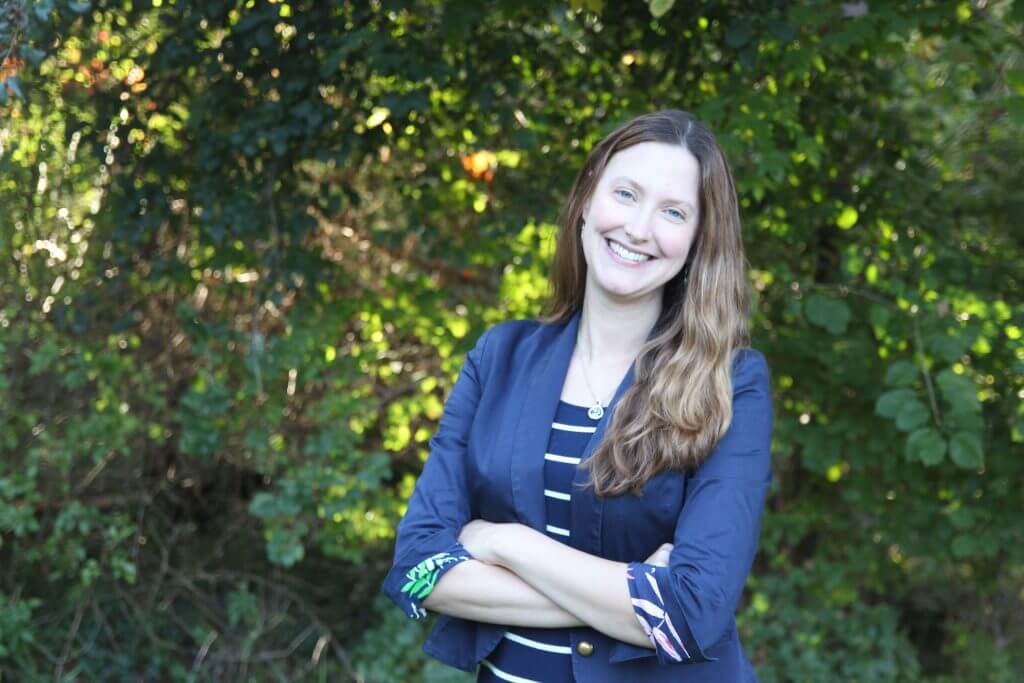 Shannon Houston, communications consultant for nonprofits and mental health providers, is standing in front of green bushes and trees, arms crossed and smiling. She is a white woman in her 30s. She is wearing a navy blue blazer over a striped shirt.