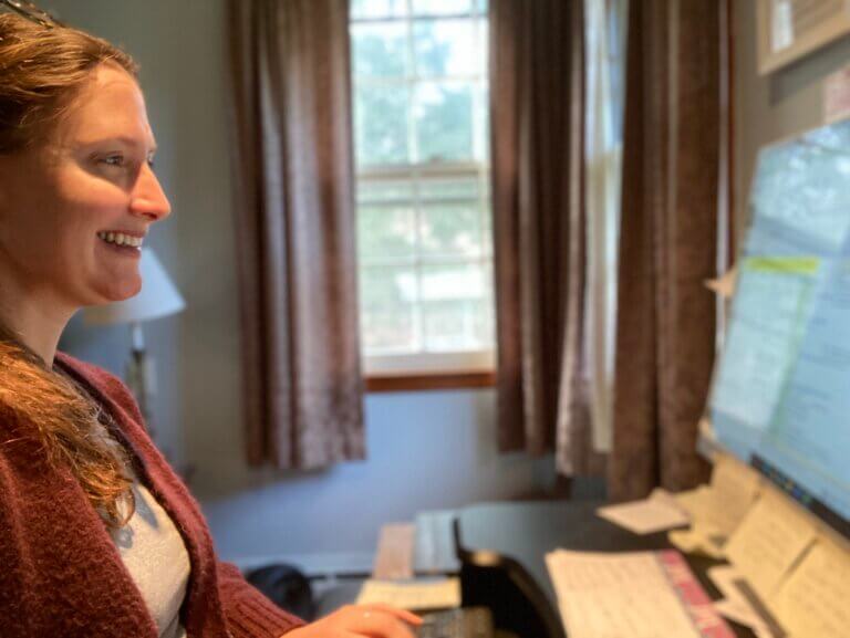 Shannon Houston, communications consultant for nonprofits and mental health providers, sits working at her computer in front of a window. The monitor has many sticky notes on it!