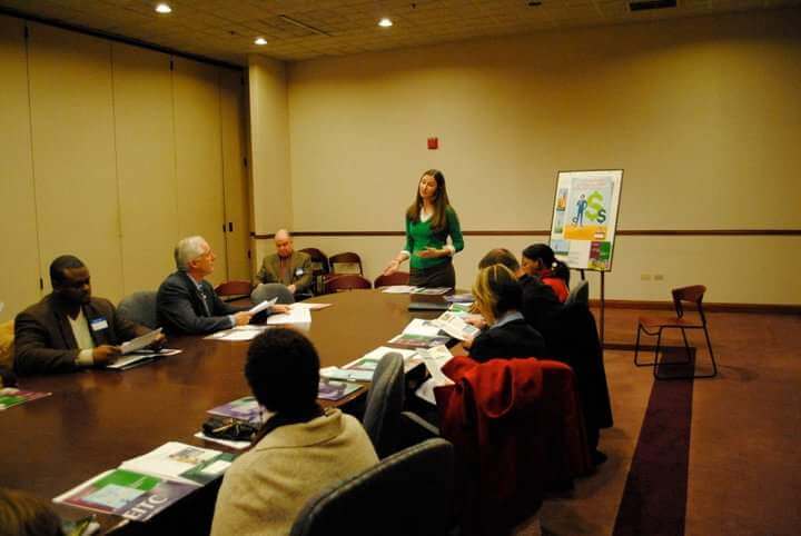 Shannon stands at the front of a long table at which seven people are seated, in what looks like a conference room. She is speaking to the group. A poster on an easel is beside her advertising a free tax program.